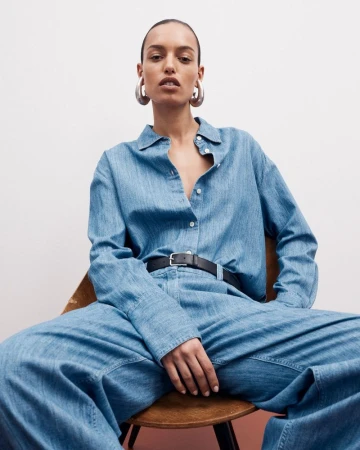 Fashion portrait of an adult woman in a denim shirt and pants, sitting in a chair against a white background, wearing silver hoop earrings.