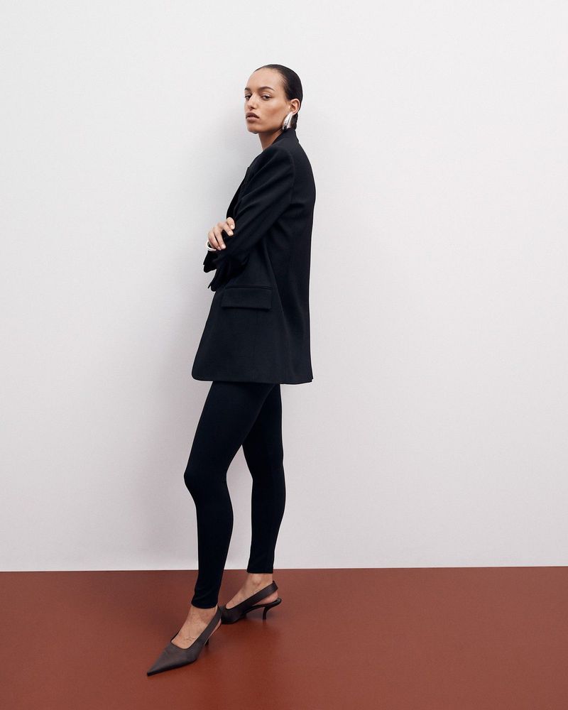 A woman in a black blazer and leggings stands in a studio with a white and brown background, creating a stylish and modern fashion shot.