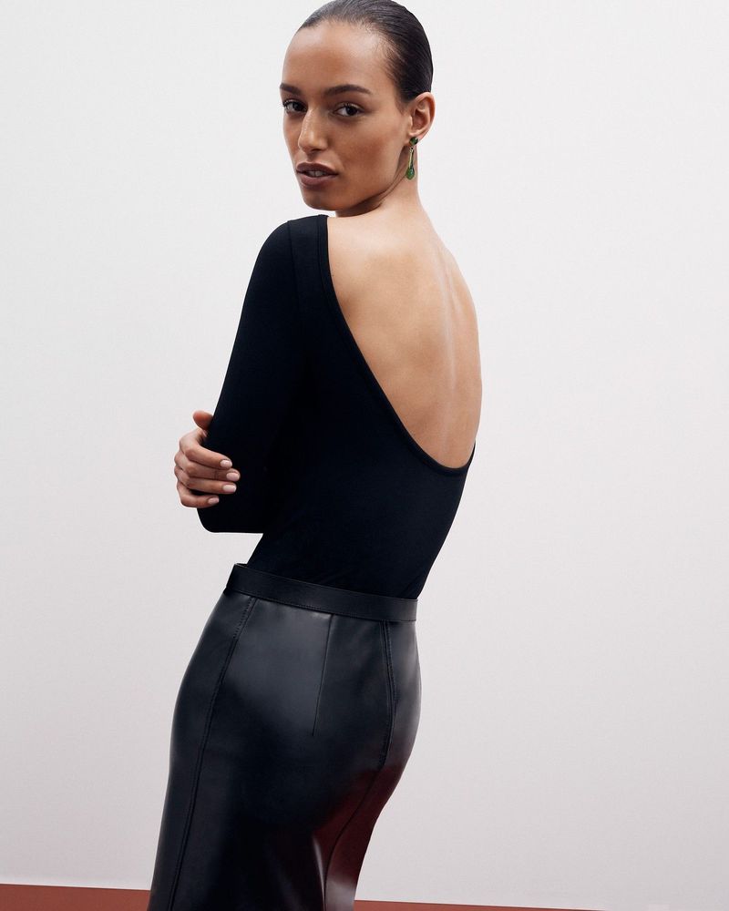 Woman posing in a black long-sleeve top and leather pencil skirt against a neutral backdrop in a fashion studio setting. 