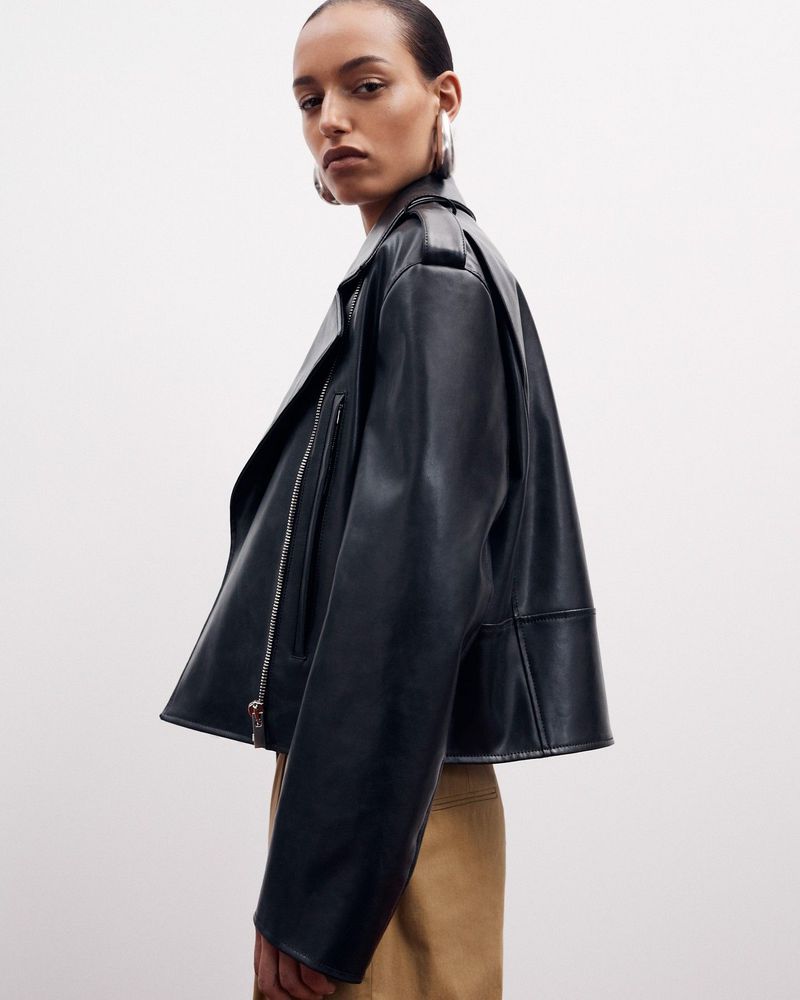 Fashion portrait of a woman wearing a black leather biker jacket and gold hoop earrings against a white studio backdrop.