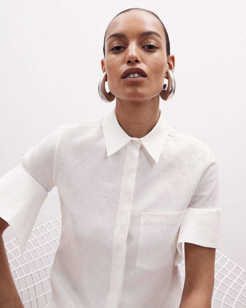 Model wearing a white button-down shirt and large silver hoop earrings seated against a white background