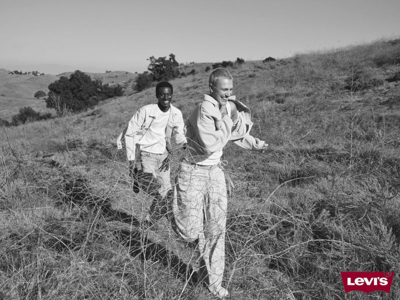 Two young men in casual clothing are running in a grassy, black-and-white landscape, with a mountain range visible in the distance.