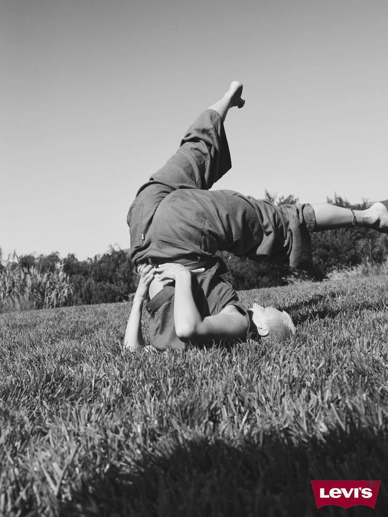 Black and white photograph of an adult doing a headstand in a grassy field with trees in the background on a bright day.