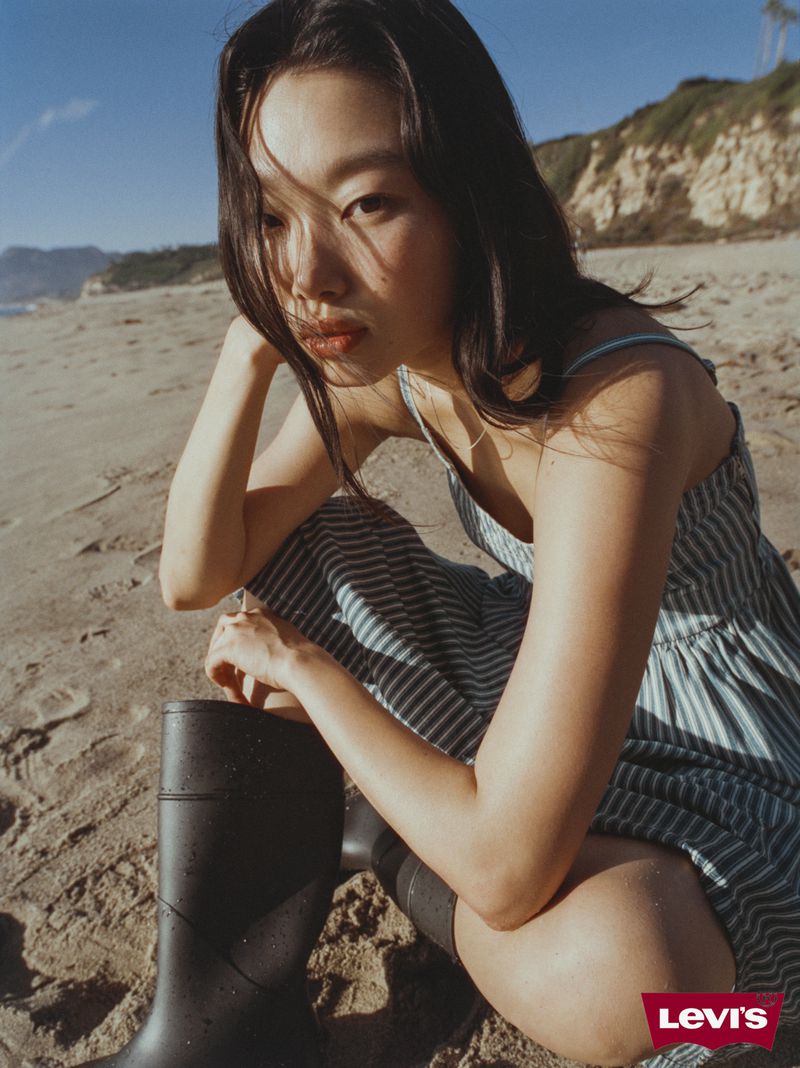 An Asian woman in a striped dress poses on a sandy beach next to rubber boots against a backdrop of blue sky and ocean.