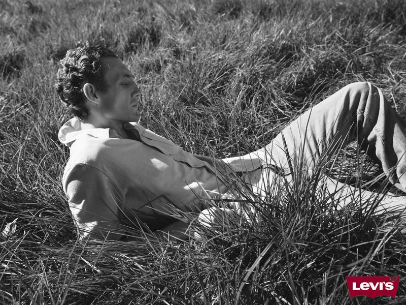 A man lying in tall meadow grass wearing denim jeans and an open shirt, photographed in black and white by Julia Noni for Levi's.