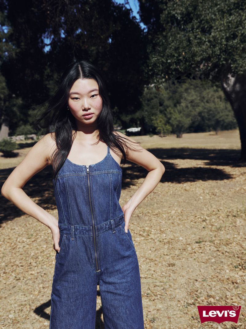 A slender Asian woman poses outdoors in a blue denim jumpsuit. She is standing in a sunny, grassy area with trees in the background.