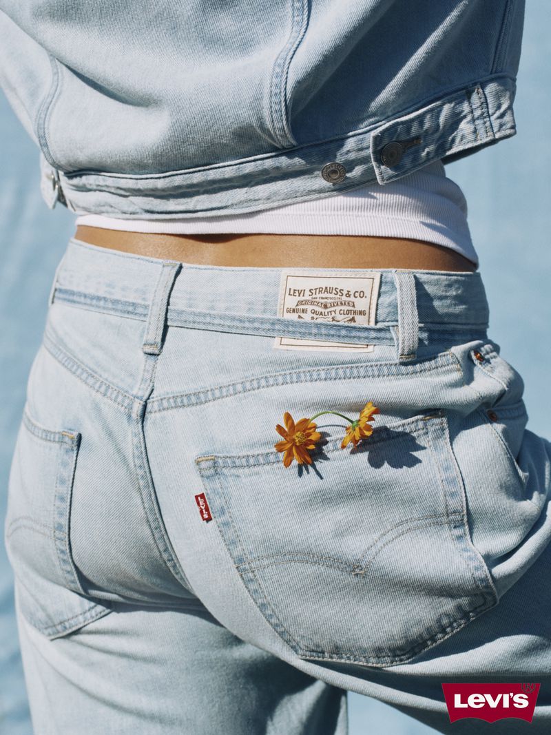 Back view of a woman wearing a light blue denim jacket and matching jeans with small orange flowers tucked into the back pocket.