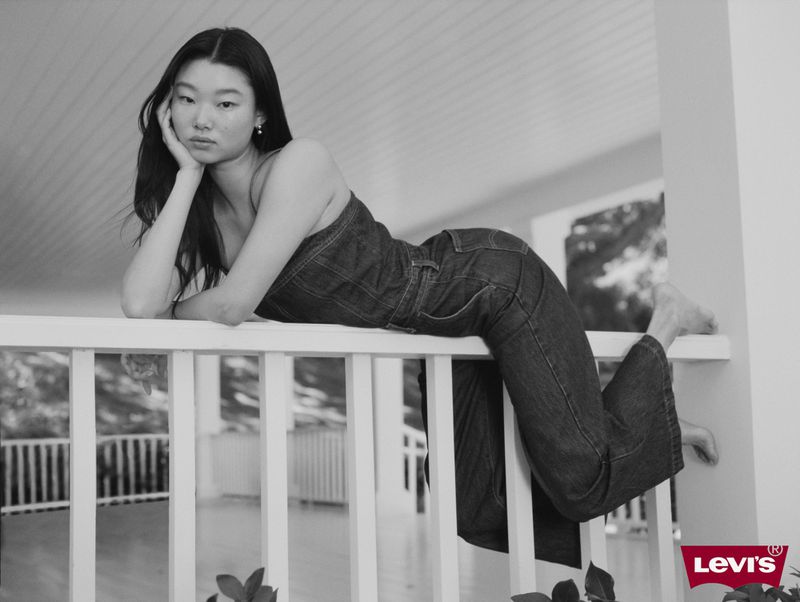 Monochrome shot of a young woman in denim outfit reclining on a white railing, with the Levi's logo visible in the lower right corner.