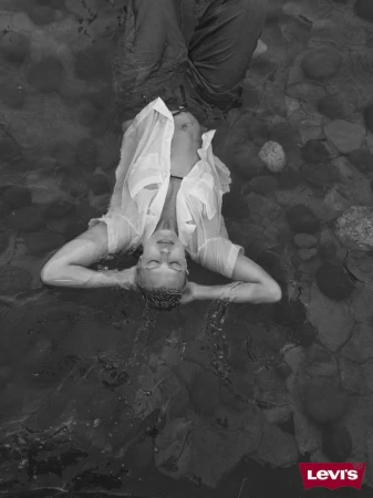 Monochrome portrait of a woman in wet shirt and jeans, lying in shallow water with rocks and the Levi's logo at the bottom.
