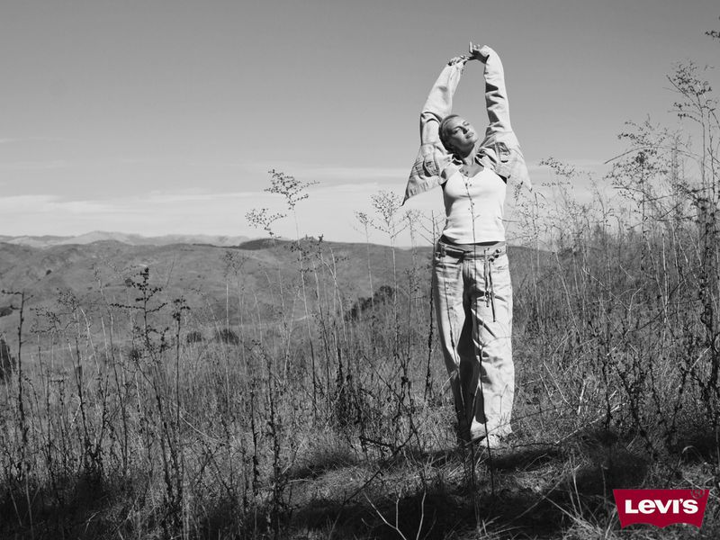 A woman stands with arms raised in a field on a hillside, wearing light-colored pants and a shirt, under a partly cloudy sky in black and white.