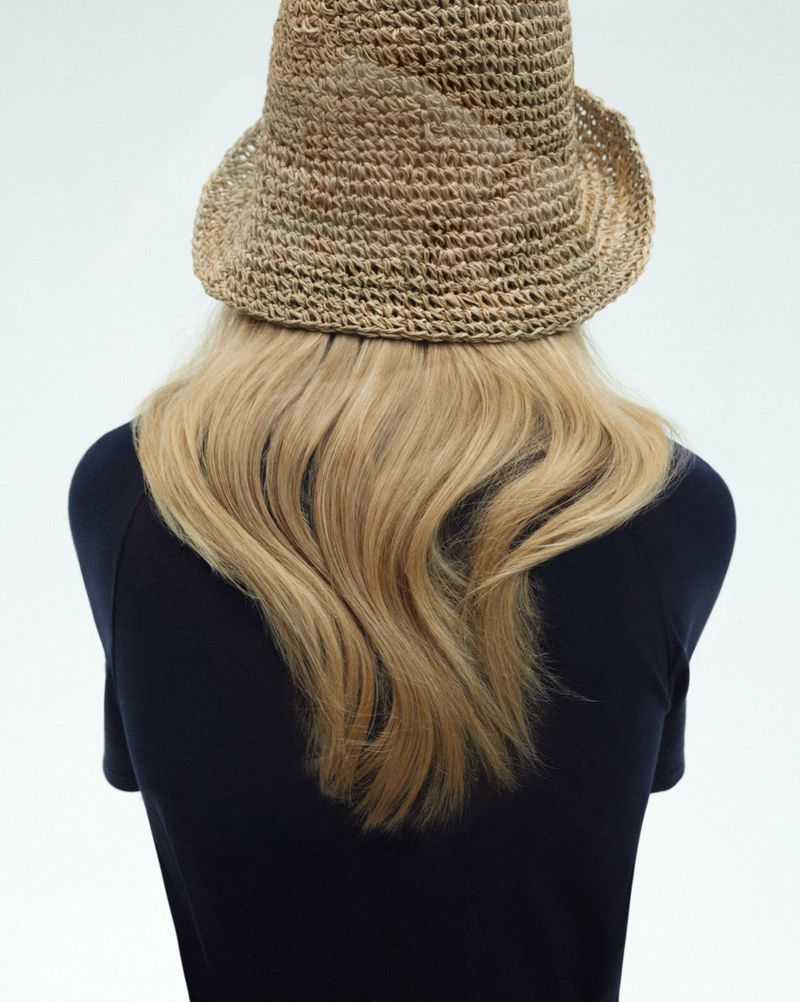 Back view of a woman with long blonde hair wearing a straw hat against a white backdrop, focusing on texture and neutral tones.
