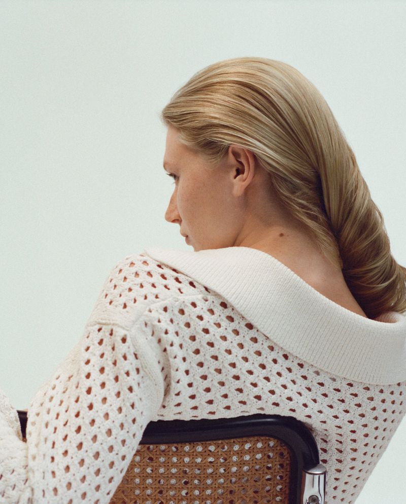 Woman with blonde hair in a twisted style, wearing an off-white knit sweater on a cane chair against a light background in soft lighting.