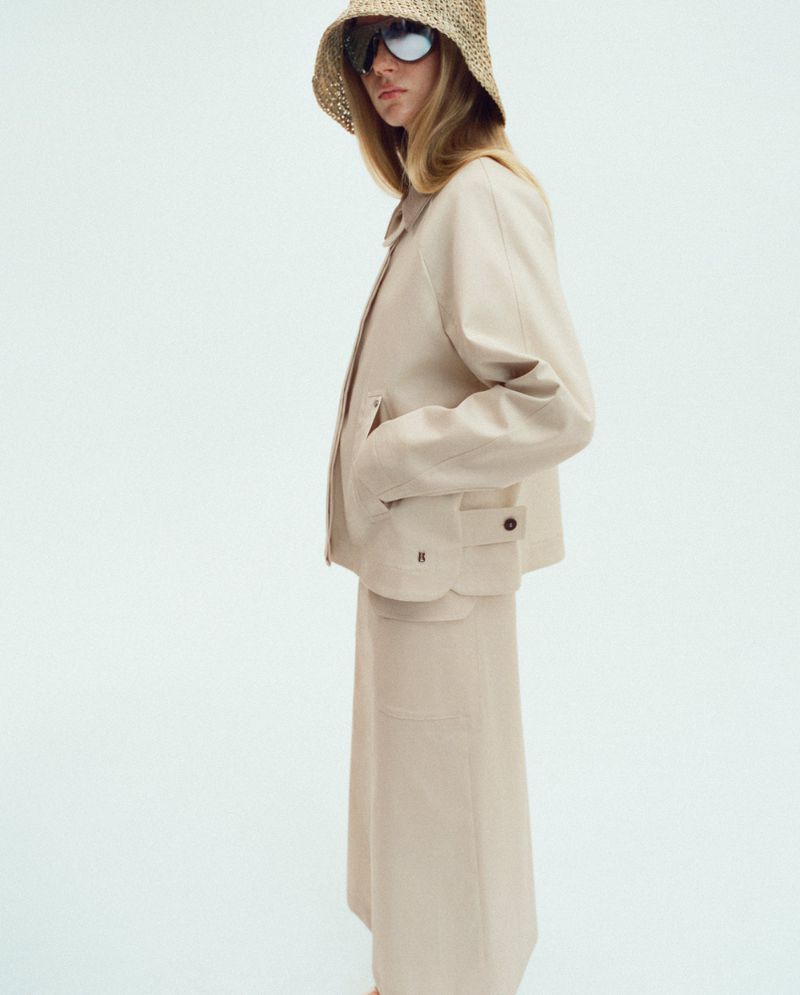 Woman in beige jacket and skirt with straw hat and sunglasses posing against white background in a fashion studio shot.