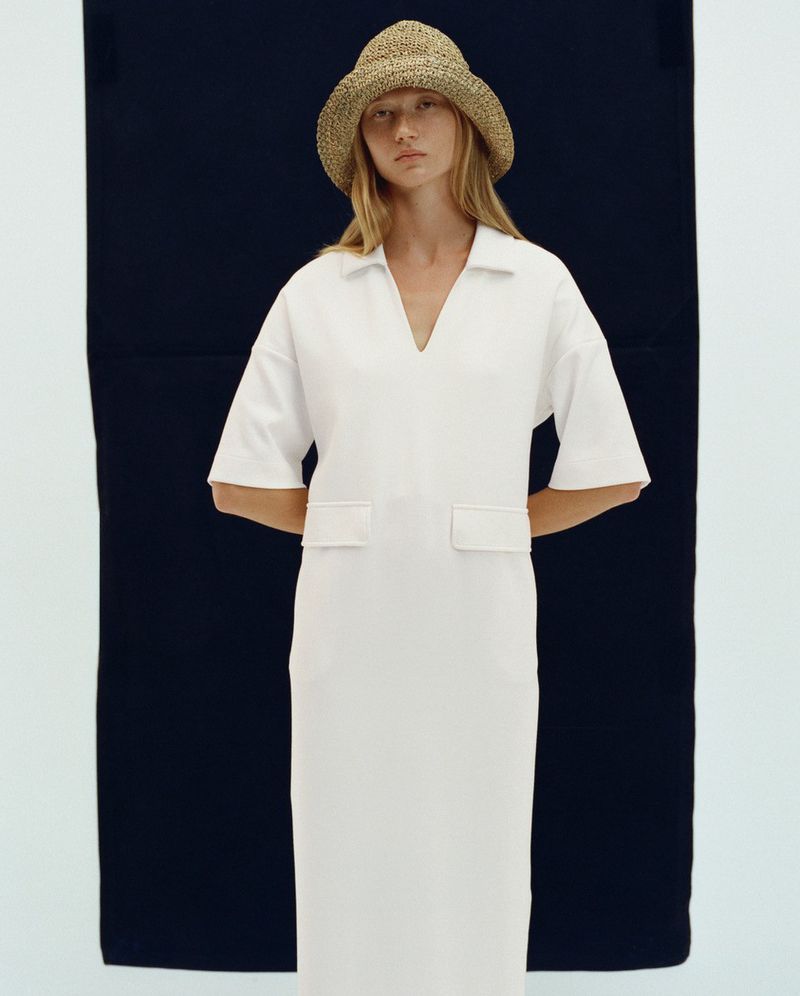 Model in a white polo dress and straw hat against a dark vertical backdrop for Bogner