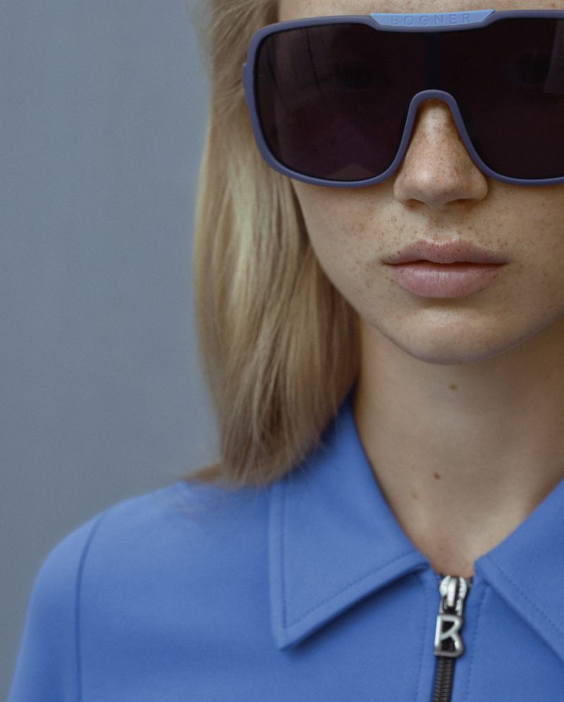 Close-up studio portrait of a blonde woman wearing oversized sunglasses, with a blue shirt and neutral grey background.