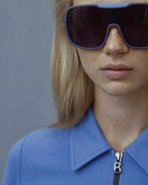 Close-up studio portrait of a blonde woman wearing oversized sunglasses, with a blue shirt and neutral grey background.