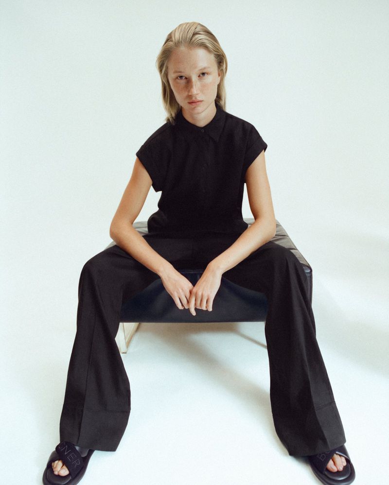 A blonde woman is seated in a photo studio wearing all black. She wears a button-up top, wide leg pants, and sandals while looking directly at the camera.