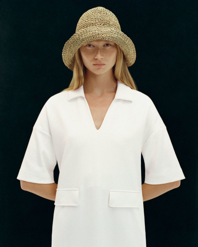 Woman in white short sleeve V-neck dress and straw hat poses in front of a dark background in a studio setting.