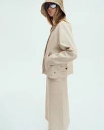 Model in a cream jacket and skirt wearing a woven bucket hat and sunglasses against a seamless white background