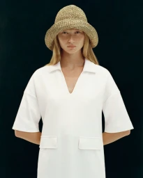 Model wearing a textured straw bucket hat and white polo dress against a dark background for Bogner