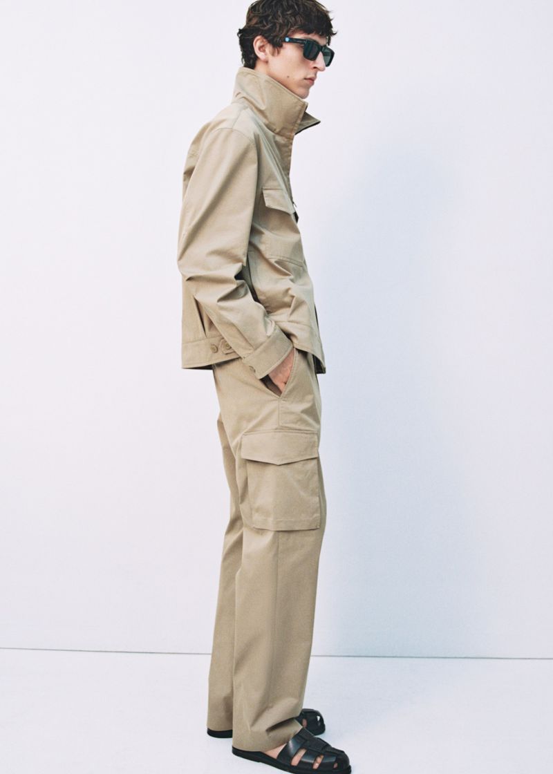A male model poses in a tan collared jacket with cargo pants and black sandals against a neutral backdrop, showcasing a fashion editorial style.
