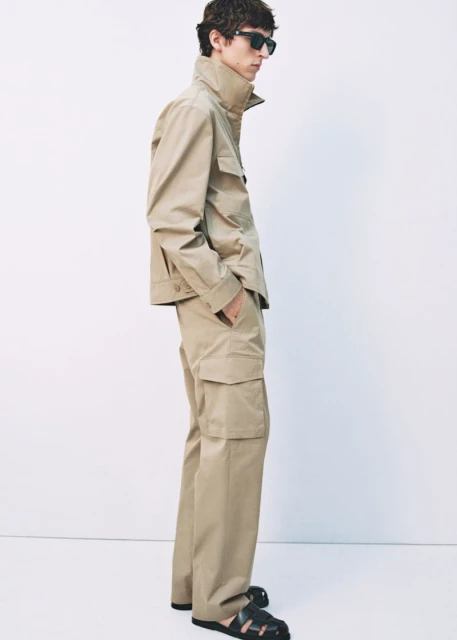 A male model poses in a tan collared jacket with cargo pants and black sandals against a neutral backdrop, showcasing a fashion editorial style.