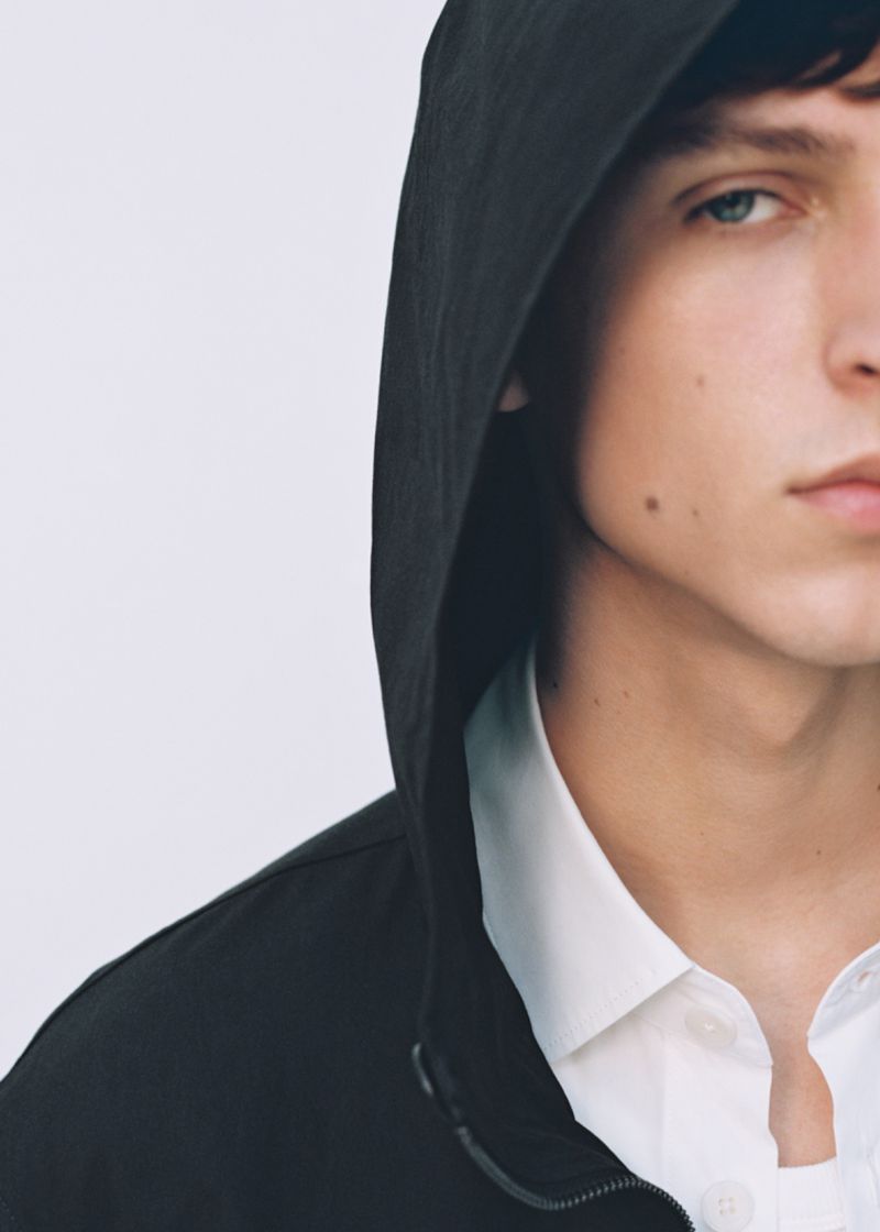 Close-up studio shot of a young adult male with blue eyes wearing a black hooded jacket over a white collared shirt against a white backdrop.
