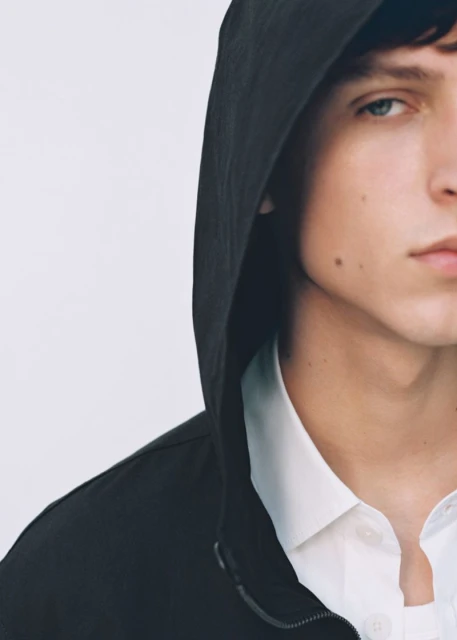 Close-up studio shot of a young adult male with blue eyes wearing a black hooded jacket over a white collared shirt against a white backdrop.