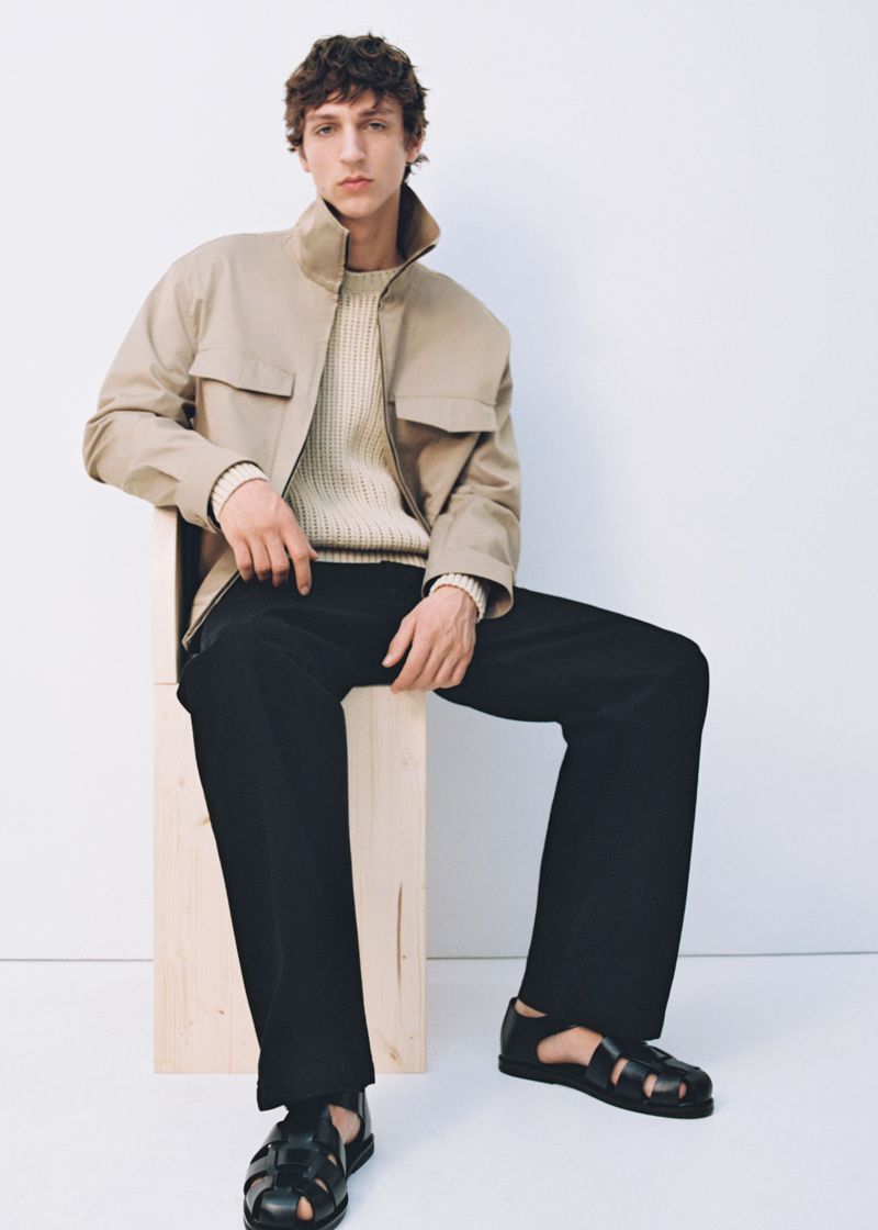 Fashion shot of an adult male model sitting on a wooden block wearing a tan jacket, light sweater, black pants, and black sandals.