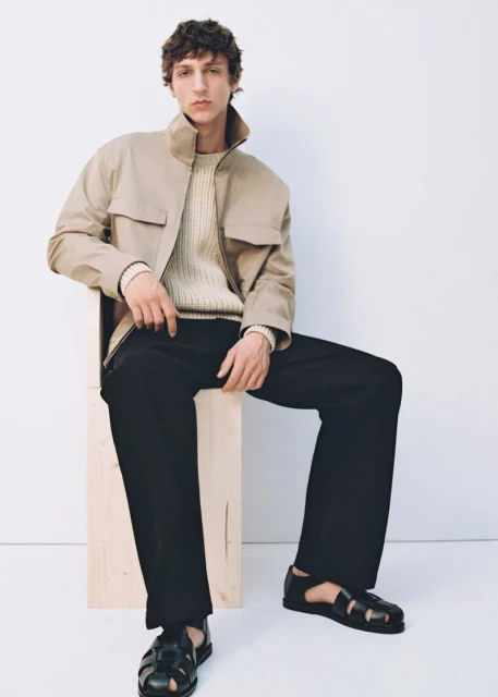 Fashion shot of an adult male model sitting on a wooden block wearing a tan jacket, light sweater, black pants, and black sandals.