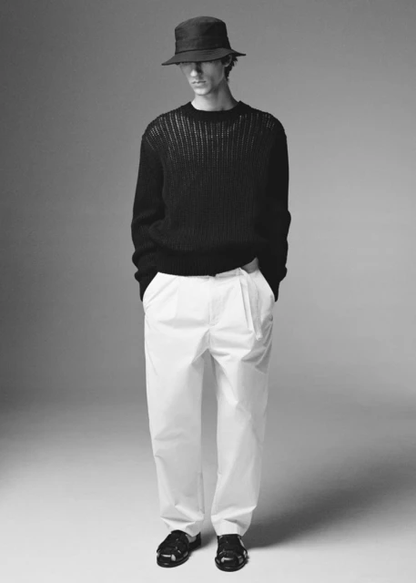 Full-length studio shot of an adult male in a dark sweater, white trousers, black shoes, and a bucket hat, standing against a grey backdrop.