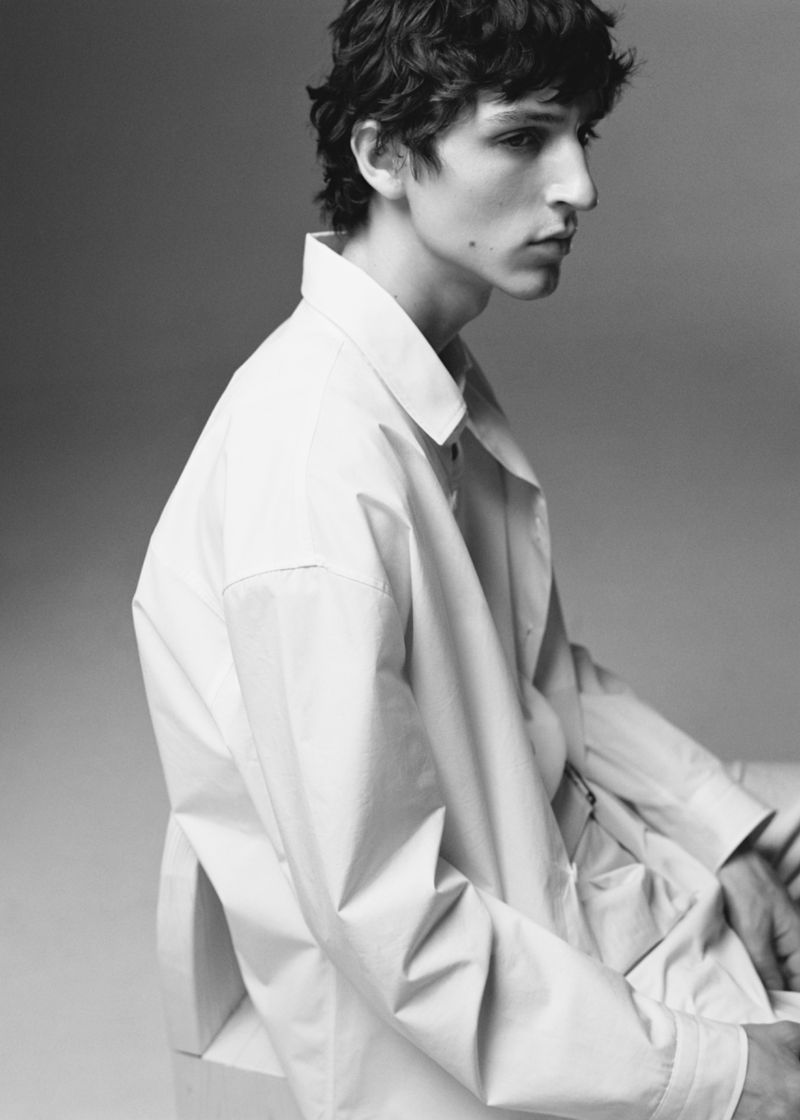 Monochrome portrait of a young male model wearing a light-coloured button-down shirt against a plain grey backdrop.