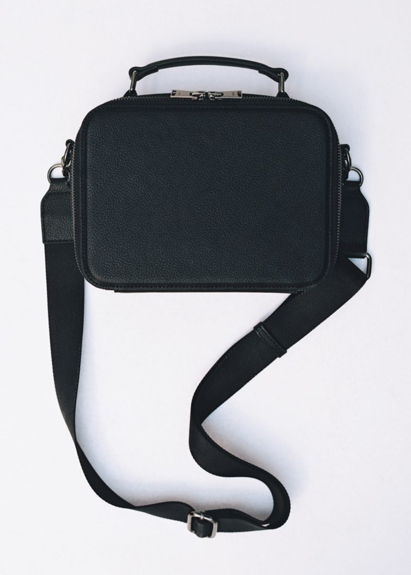 Square-shaped black leather bag with handle and strap, laid flat on a white surface in a studio setting with even lighting.
