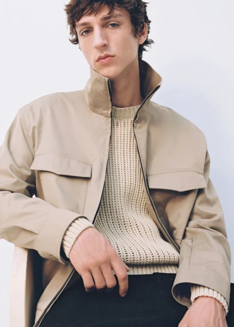 Adult male poses indoors wearing a beige jacket over a cream-colored sweater, complemented by black pants. Neutral backdrop.
