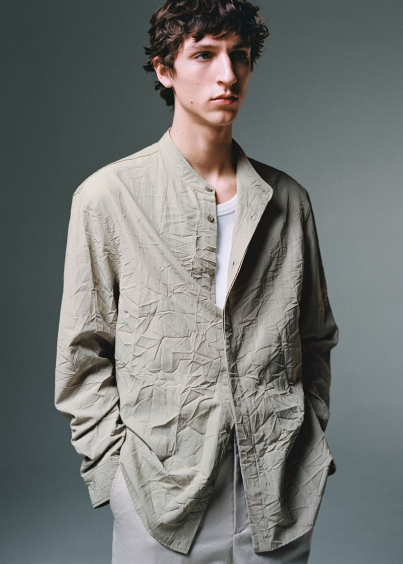 A young male model with dark wavy hair wearing a textured light grey button-down shirt over a white undershirt against a neutral grey studio backdrop.