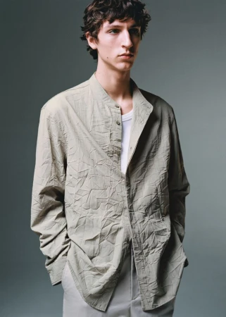 A young male model with dark wavy hair wearing a textured light grey button-down shirt over a white undershirt against a neutral grey studio backdrop.