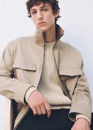 A young male model wearing a beige utility jacket over a cream knit sweater against a plain white studio background.