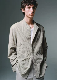 A young male model with dark wavy hair wearing a textured light grey button-down shirt over a white undershirt against a neutral grey studio backdrop.