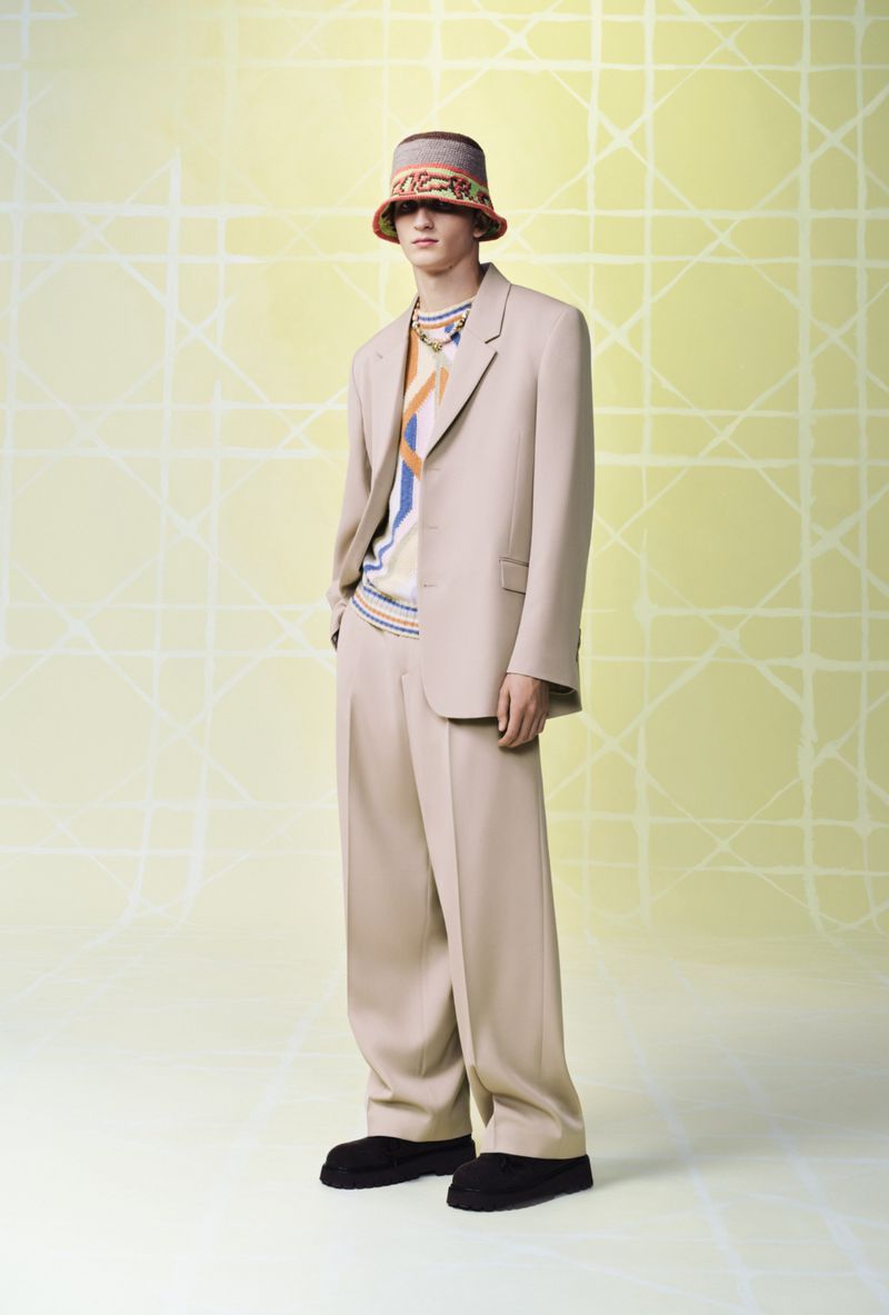 A slender male model poses in a tan suit and patterned bucket hat against a yellow background, showcasing high fashion.