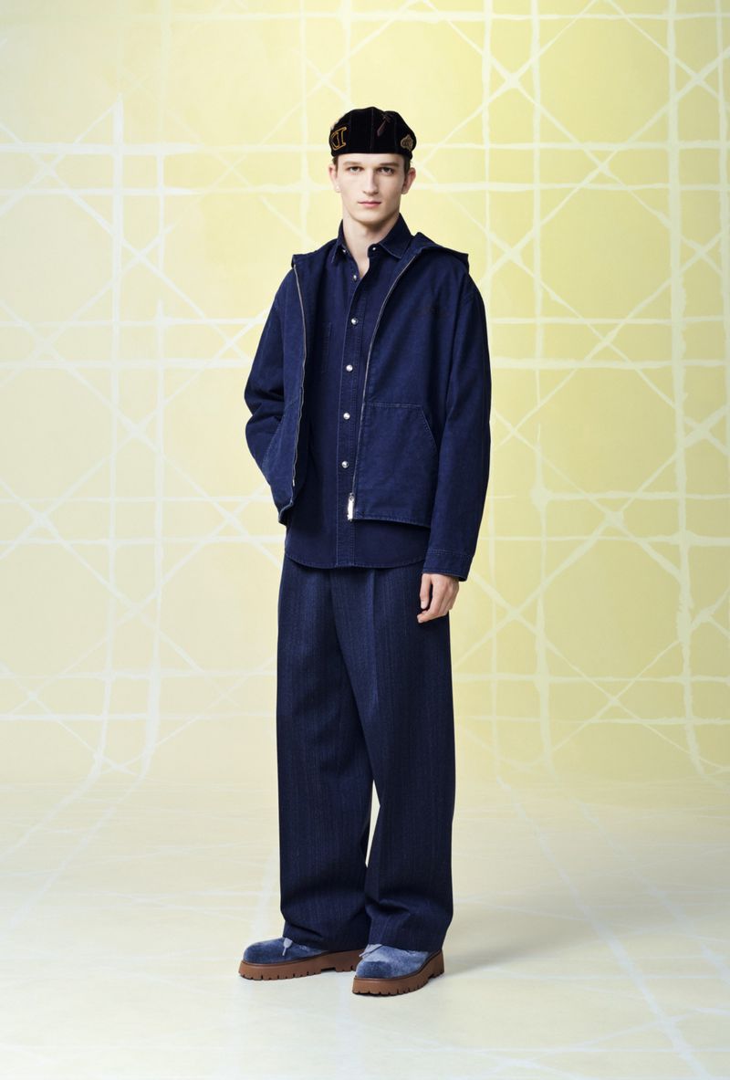 Full shot of a young adult male fashion model posing in dark blue denim clothing and a black cap against a geometric pale yellow backdrop.