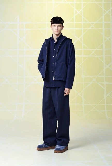 Full shot of a young adult male fashion model posing in dark blue denim clothing and a black cap against a geometric pale yellow backdrop.