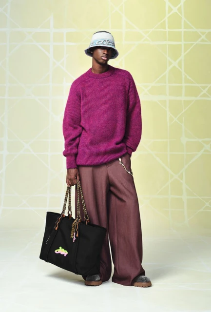 Full shot of an adult male model wearing a magenta sweater, wide-legged brown pants, and a bucket hat, holding a black tote bag.