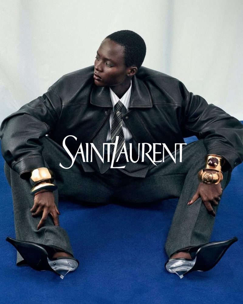 Fashion shot of a woman in a leather jacket, gray pants, and black high heels, posing on a blue surface with the SAINT LAURENT logo.