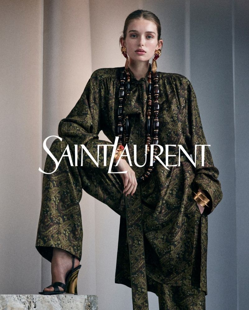 A female model showcases Saint Laurent apparel, donning a patterned top and pants, paired with jewelry, while posing indoors.