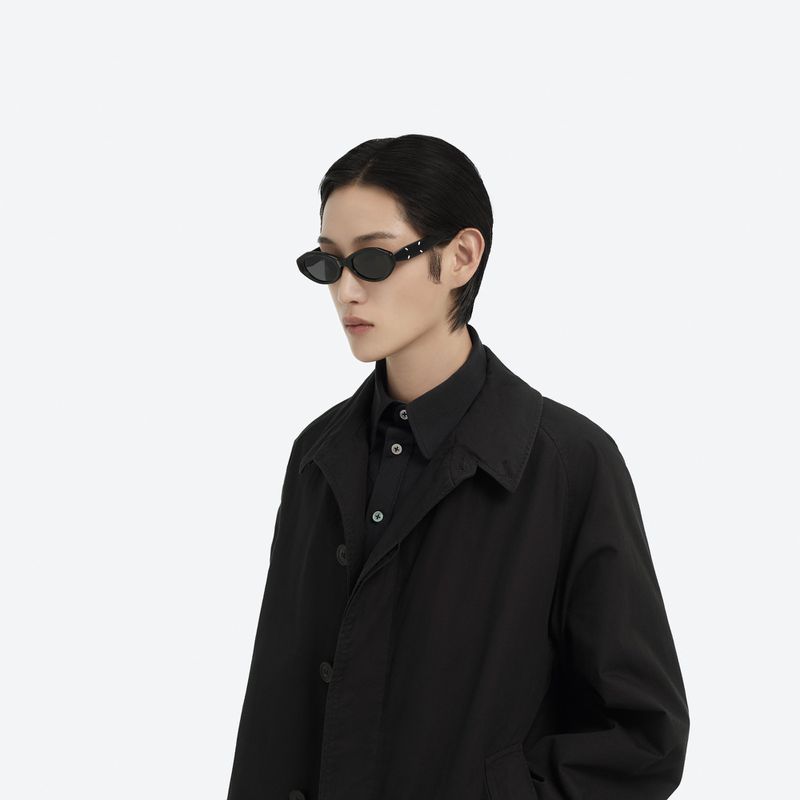 A person with short hair and wearing sunglasses and a black jacket stands against a white background, showcasing fashion and style.