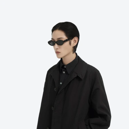 A person with short hair and wearing sunglasses and a black jacket stands against a white background, showcasing fashion and style.