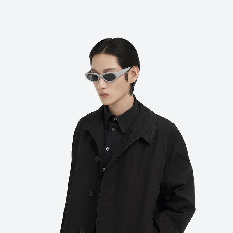 Portrait of a person with black hair and wearing sunglasses and a black trench coat, against a plain white background in a studio.