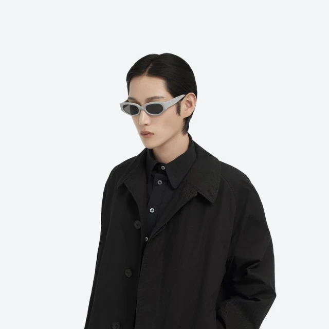 Portrait of a person with black hair and wearing sunglasses and a black trench coat, against a plain white background in a studio.