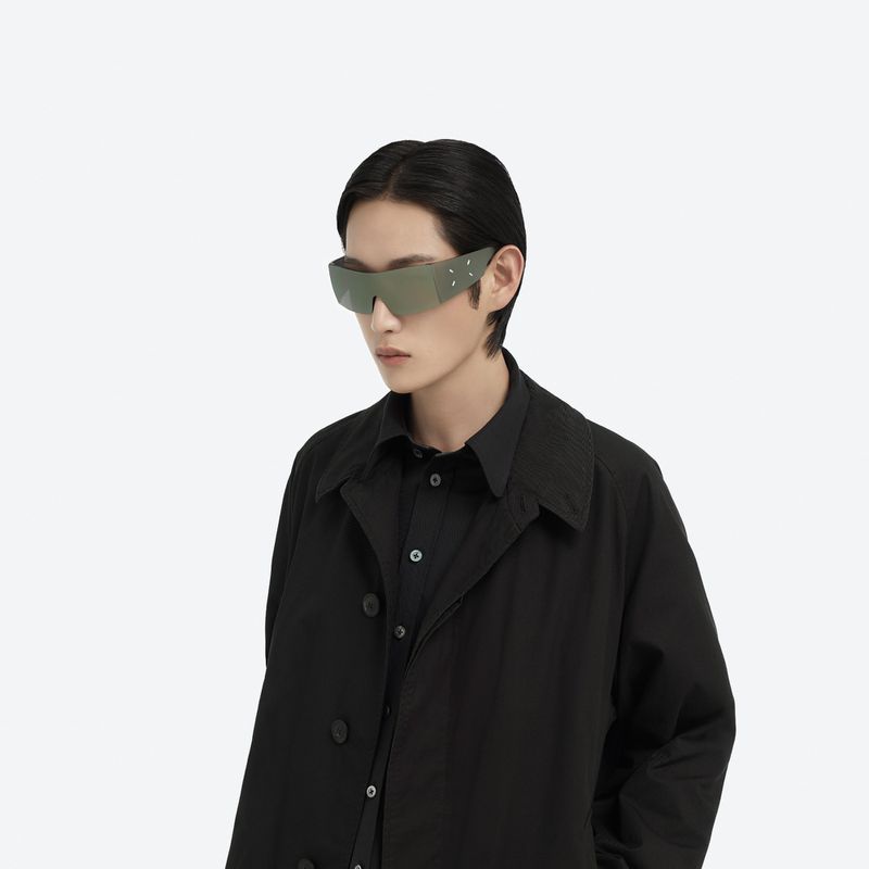 Model wearing futuristic wraparound sunglasses and black coat against a white background for Maison Margiela X Gentle Monster
