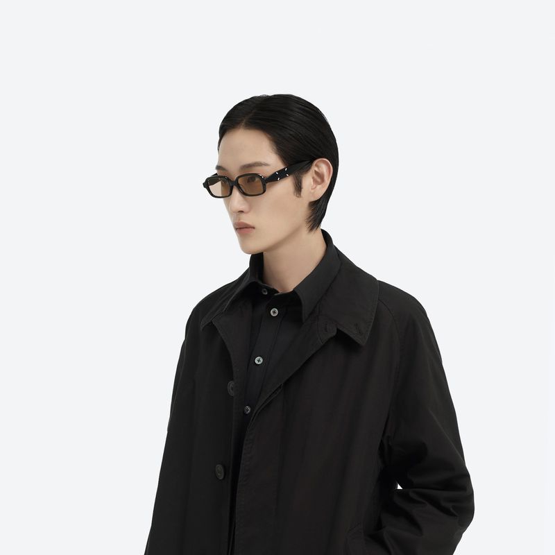 Close-up studio shot of an adult with dark hair wearing a black shirt and dark coat with a pair of retro framed glasses.